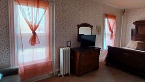 Ein Zimmer mit gestreifter Tapete, einem Heizkörper, einem Holzschrank und einem Fernseher darauf sowie einem Bett mit Kopfteil.