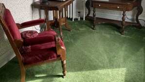 Une pièce avec un fauteuil rouge, une petite table avec une lampe, un téléviseur posé sur un meuble en bois et une fenêtre avec des stores.