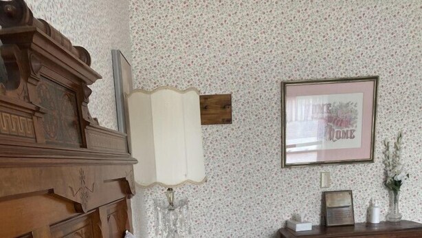 Une chambre à coucher avec une tête de lit en bois, une courtepointe à motifs floraux, un oreiller de couleur crème, une table de chevet avec une lampe et un tableau encadré accroché au mur.