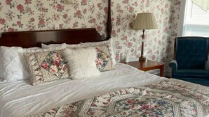 A bedroom with a large bed, floral wallpaper, a wooden headboard, a nightstand, a lamp, and a chair. - AH #28 · AH #28 King w/Jacuzzi/SS/Fridge/Micro (Wabasha)