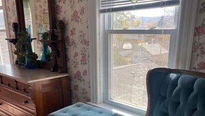 A room with a large window, a blue upholstered armchair, a wooden dresser, and a mirror with floral wallpaper. - AH #28 · AH #28 King w/Jacuzzi/SS/Fridge/Micro (Wabasha)