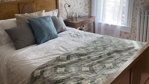 Une chambre à coucher avec un lit en bois, un papier peint à motifs floraux, un tableau encadré, une table de chevet avec une lampe et une fenêtre avec un rideau vert.