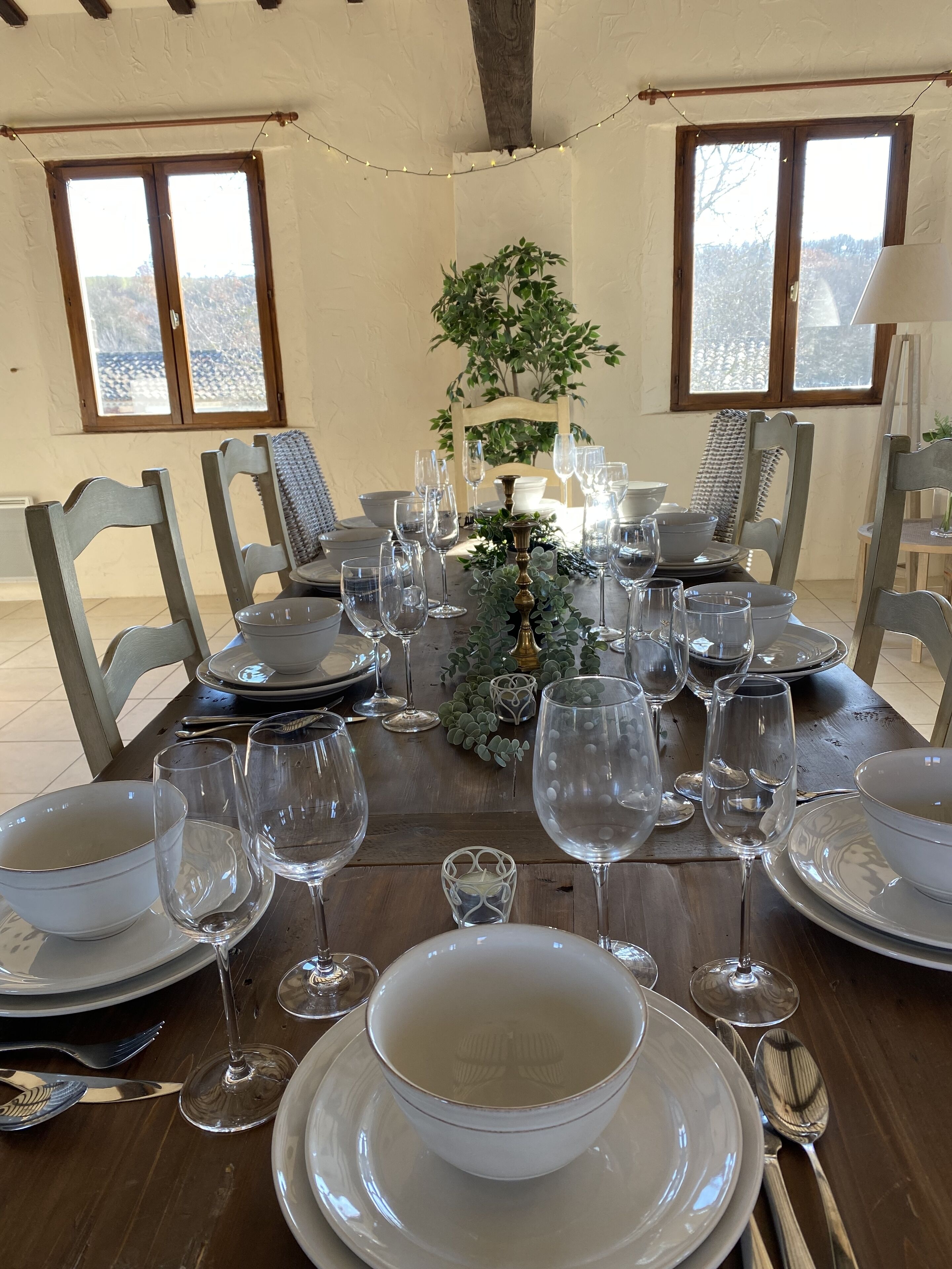Een eettafel gedekt met witte borden, kommen en wijnglazen, omringd door stoelen, met een plant als middelpunt en lichtslingers.