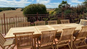 Un patio al aire libre con una mesa y sillas de madera, rodeado por una cerca de metal, con vistas a un paisaje pintoresco.