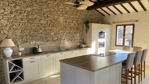 Una cocina con pared de piedra, vigas de madera, una isla central y una zona de comedor con sillas.