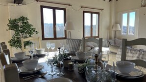 Un comedor con una mesa de madera dispuesta para una comida, rodeado de sillas, un sofá y dos ventanas con cortinas.