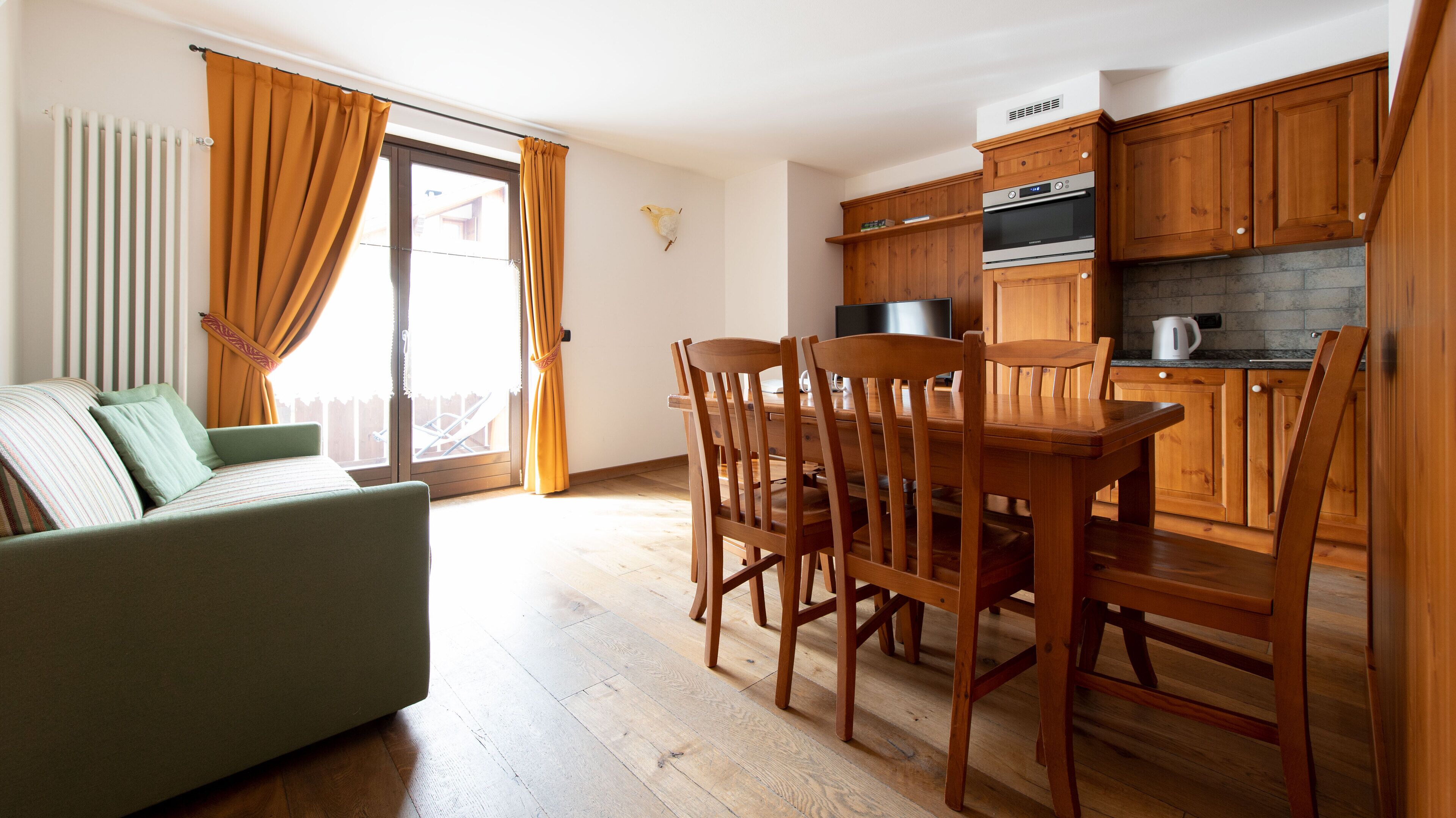 Un salón acogedor con sofá, una mesa de comedor de madera y sillas. Se ve una cocina con armarios de madera y horno empotrado.