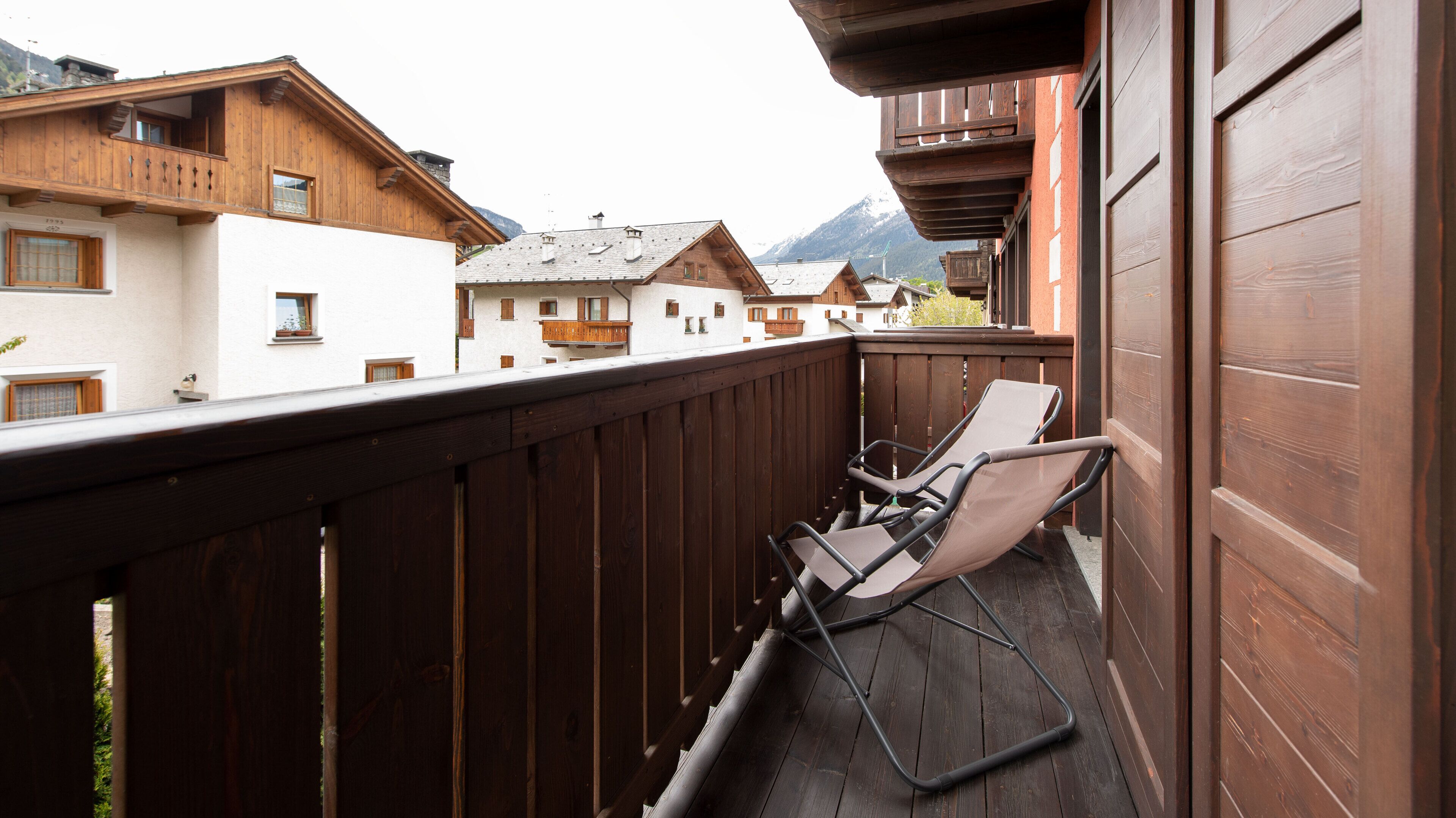 Un balcón de madera con dos sillones de descanso, una valla de madera y vistas a edificios cercanos y montañas.