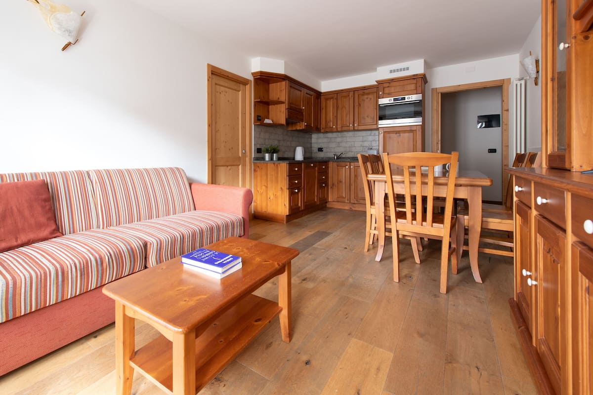 A compact living space with a sofa, a dining table with chairs, wooden cabinets, and a kitchen area with a stove.