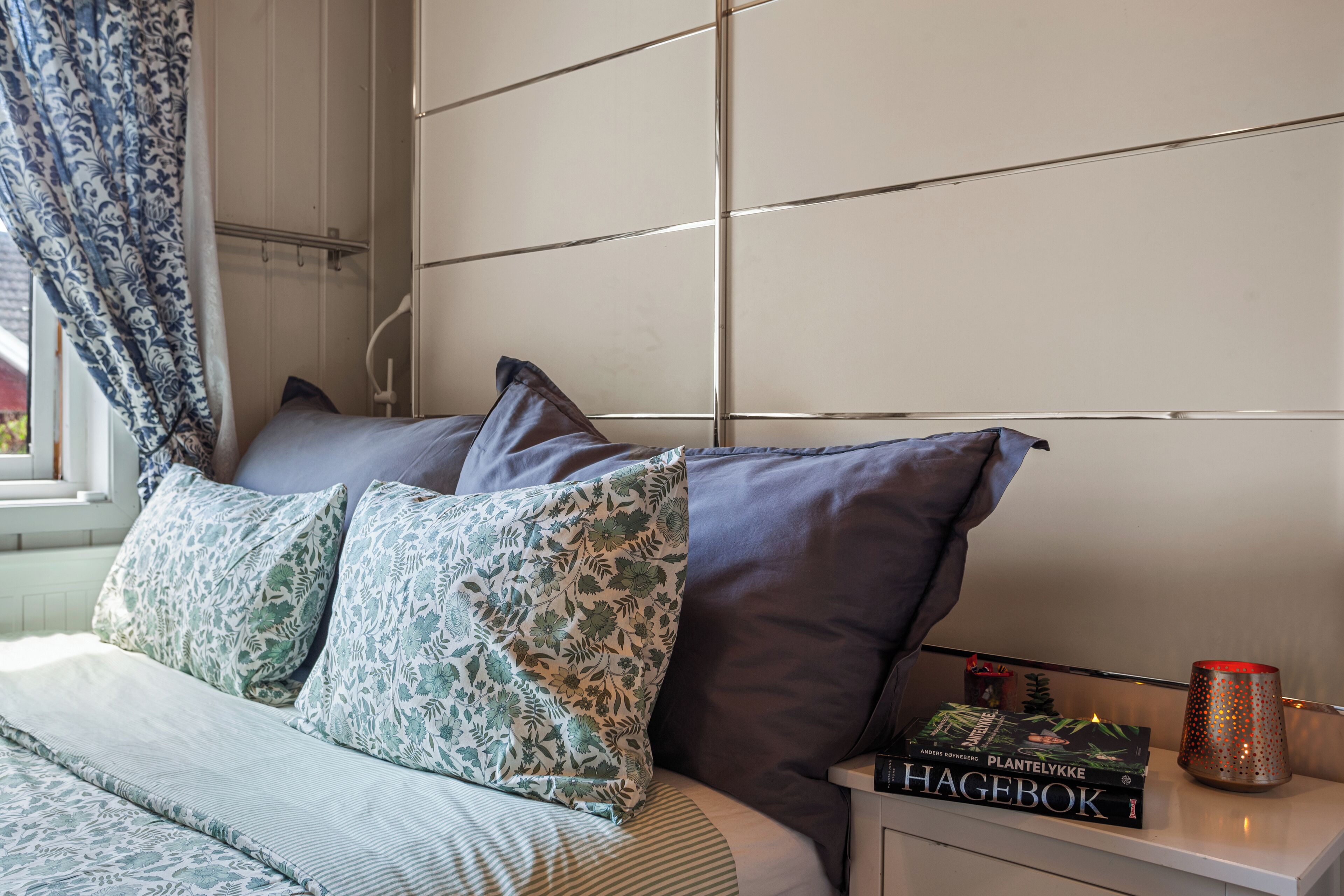 Una cama bien hecha con almohadas de estampado floral, un libro titulado 'Hagebók' sobre una mesita de noche y una vela decorativa.