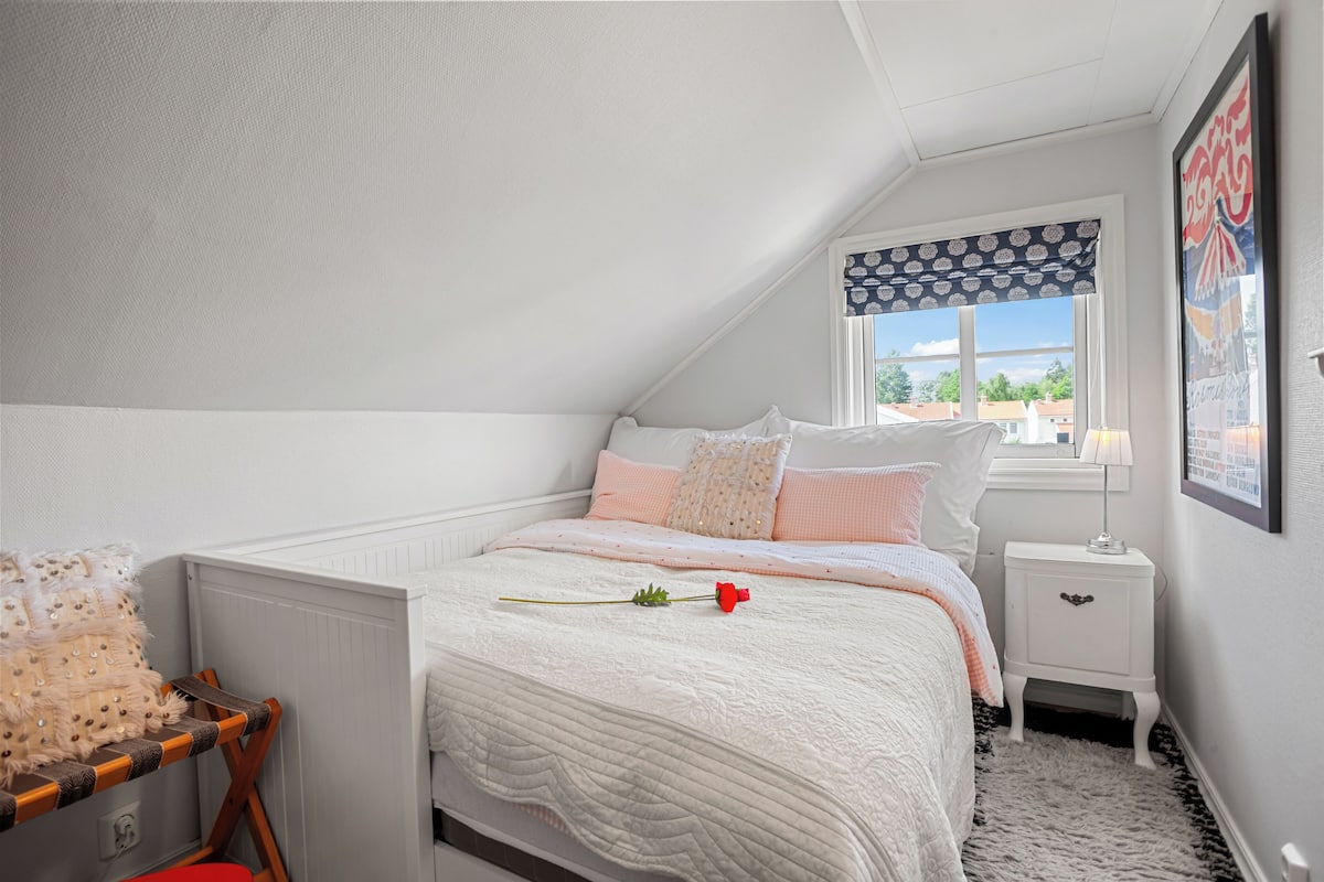 A bedroom with a bed, a nightstand, a window with patterned curtains, and a framed picture on the wall.