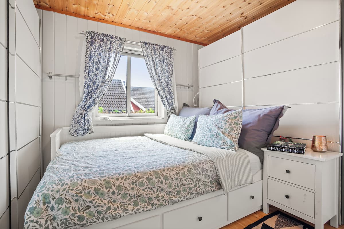 A bedroom with a bed, a nightstand, a window with curtains, and a view of a house outside.
