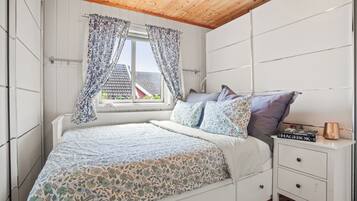 A bedroom with a bed, a nightstand, a window with curtains, and a view of a house outside.