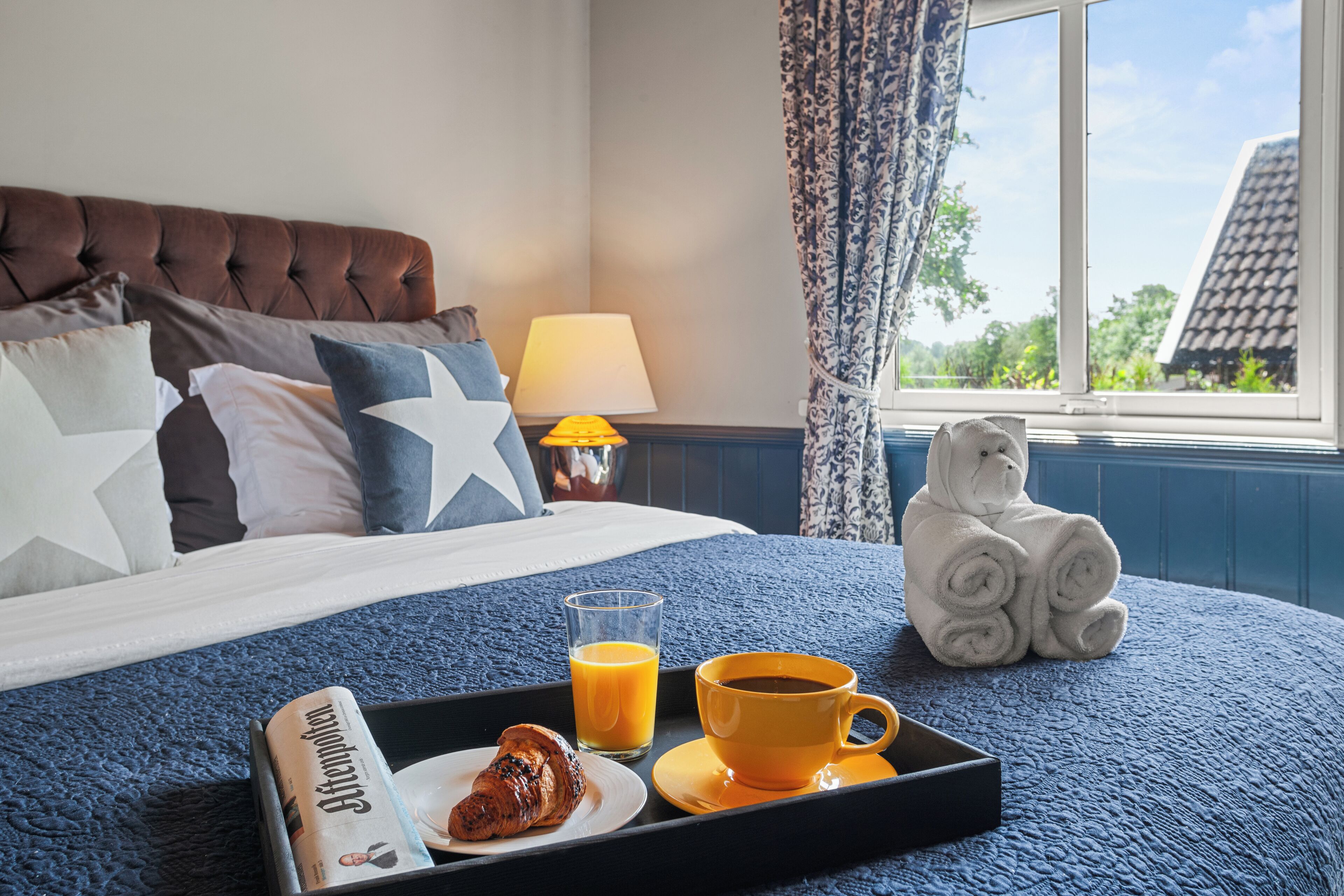 A neatly made bed with a blue blanket, pillows, a tray with croissant, orange juice, and coffee, and a stuffed animal on a window sill.