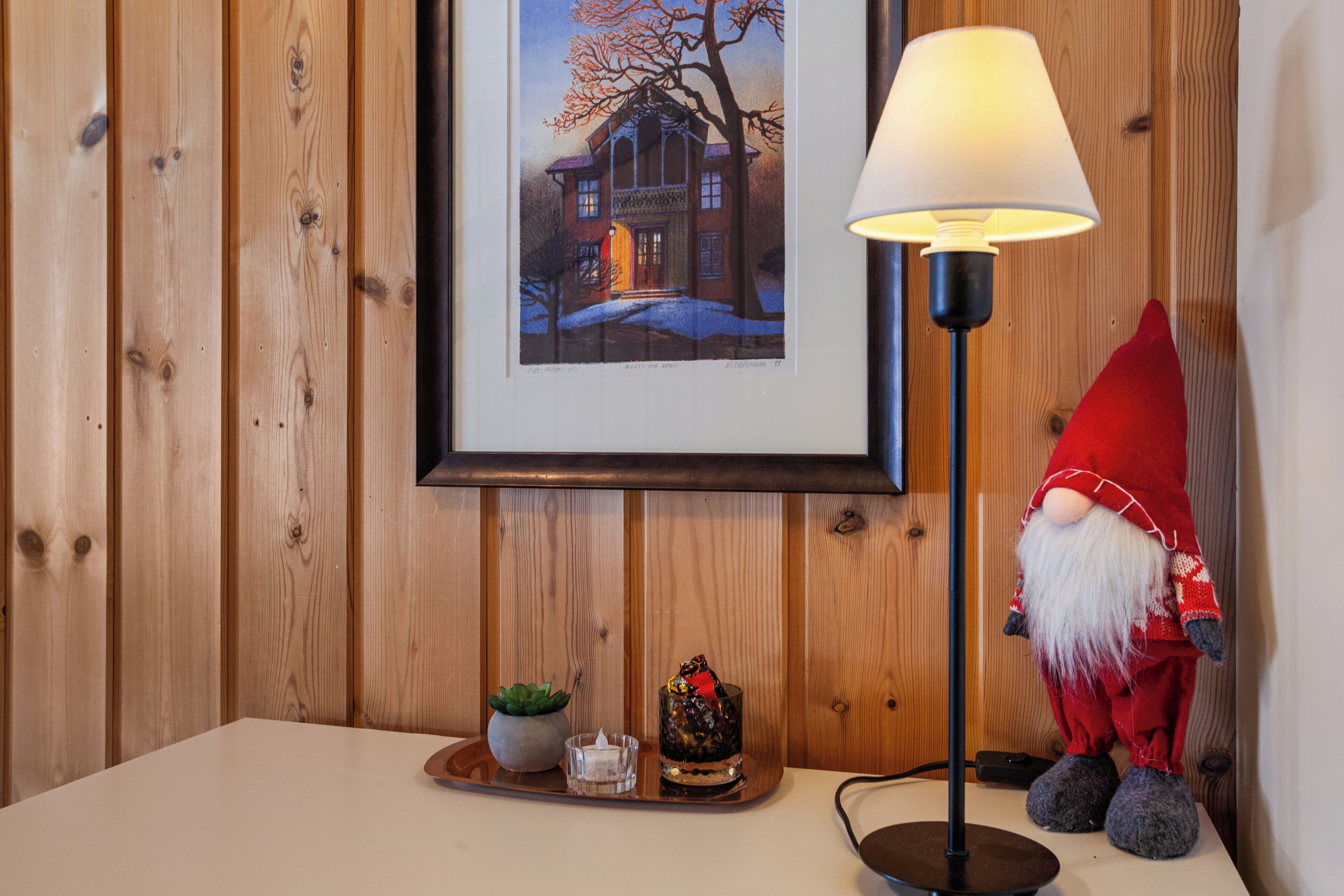 Pared de madera con un cuadro enmarcado, una lámpara de pie, un gnomo rojo y una mesita con una vela y un vaso.