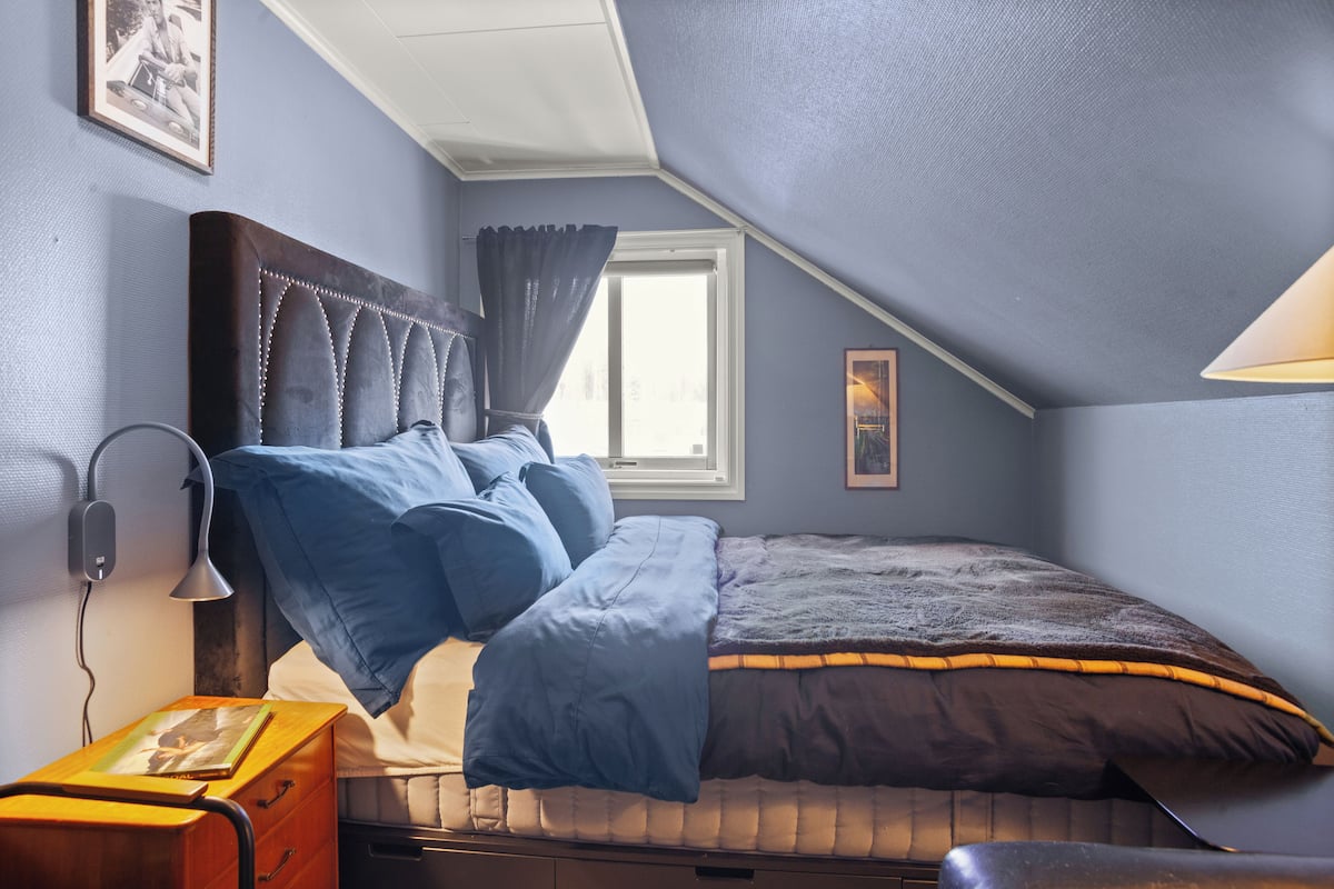 A bedroom with a bed, a nightstand, a lamp, a window with curtains, and a picture on the wall.