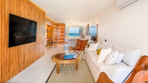 Ein modernes Wohnzimmer mit einem an der Wand befestigten Flachbildfernseher, einem runden Holztisch, einem weißen Sofa mit Kissen und Meerblick durch große Fenster.