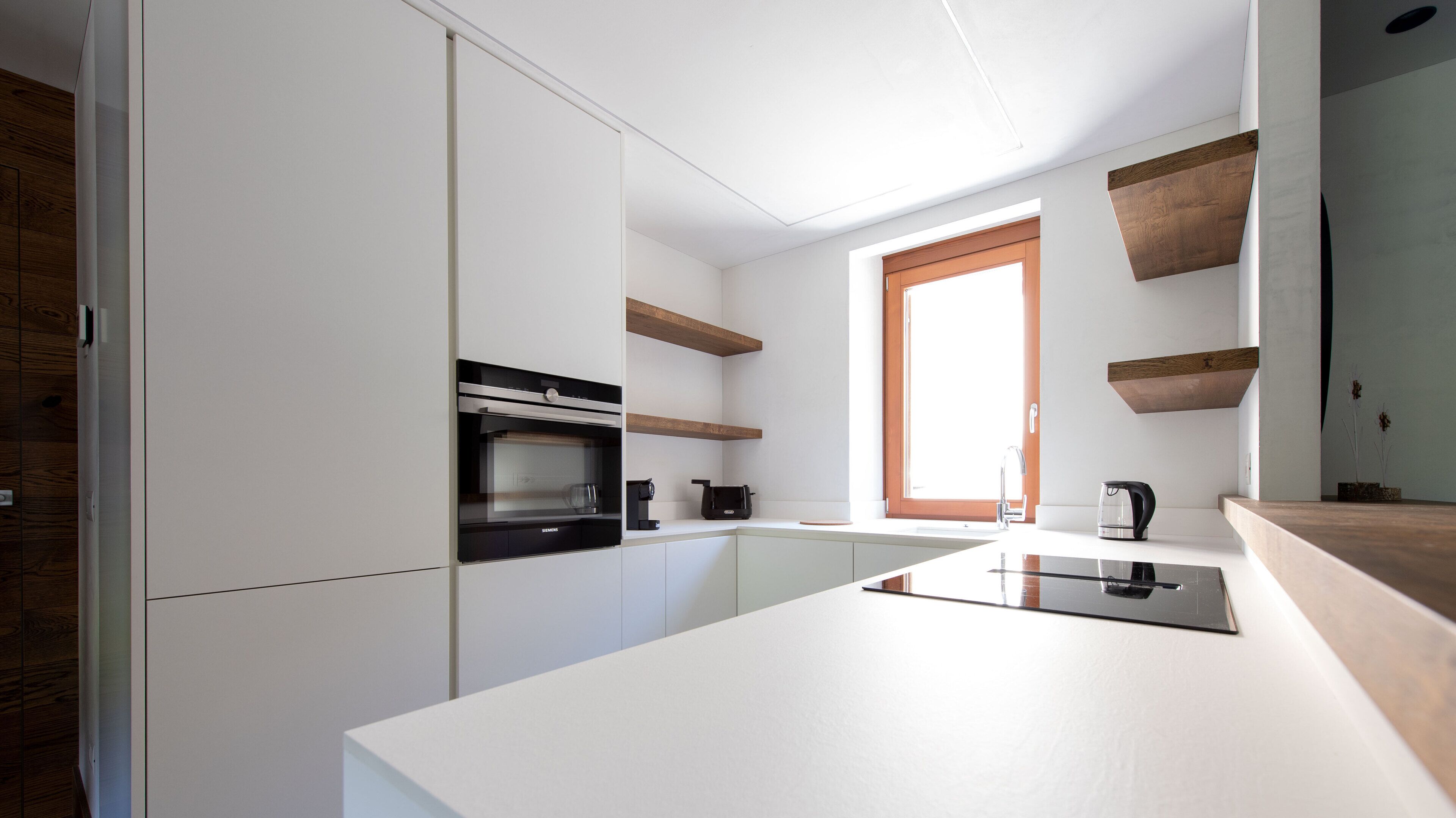 A modern kitchen with white cabinetry, a built-in oven, a coffee machine, and wooden shelves.