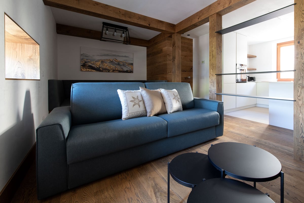 A modern living room with a blue sofa, two round tables, wooden beams, and a kitchen in the background.