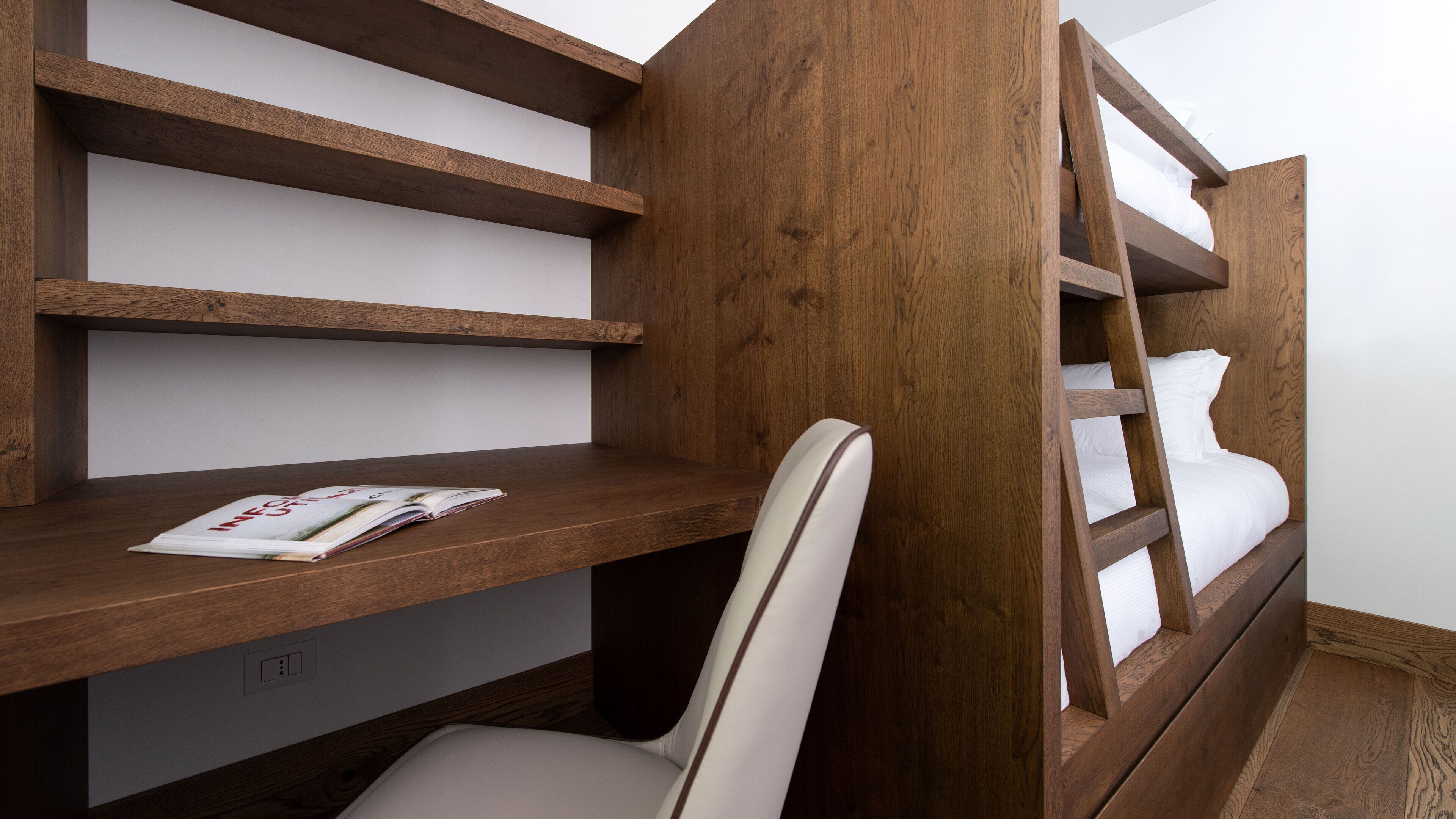 A bunk bed with wooden headboard and shelves, a white chair, and a magazine on the bed.