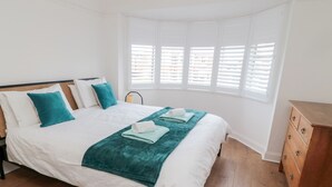 A neatly made bed with white and teal bedding, a wooden dresser, and a window with blinds. - Shileen (Seahouses)
