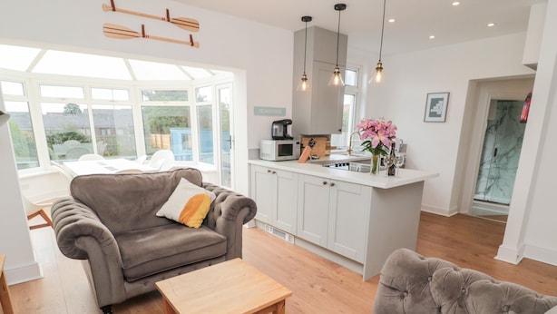 A modern living room with a grey sofa, a wooden coffee table, and a grey armchair. There is a kitchen area with white cabinets and a microwave. - Shileen (Seahouses)