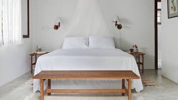 Un dormitorio con una cama, dos mesitas de noche, un banco, una lámpara de noche, una lámpara de pared y una ventana con cortinas.