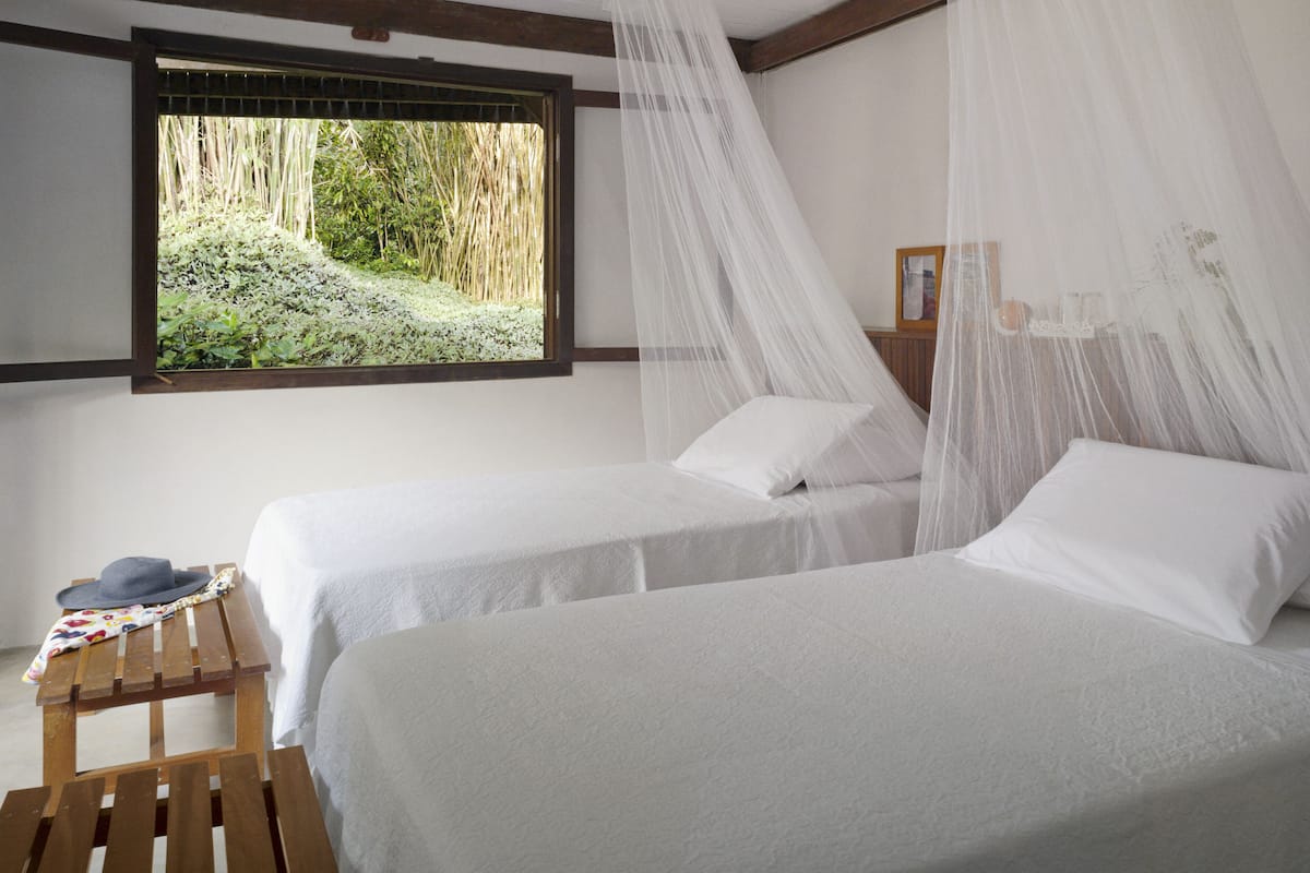 Ein Zimmer mit zwei Betten, einem Fenster mit Blick auf Grünflächen, einem Holztisch mit Hut und Blumenunterlage sowie einem Nachttisch mit Lampe.