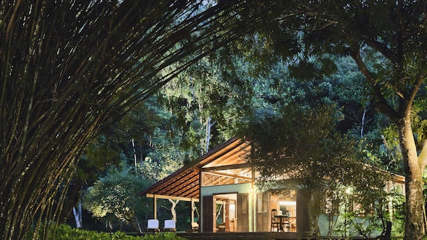 Una casa de madera con porche cubierto, rodeada de abundante vegetación y un puente de piedra.