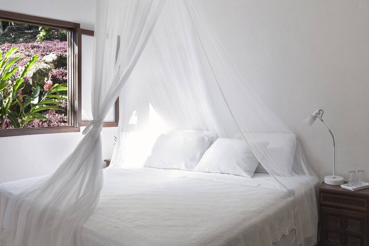 Ein Bett mit weißen Leinen und einem Baldachin, ein Nachttisch mit Lampe sowie ein Fenster mit Blick auf Pflanzen.