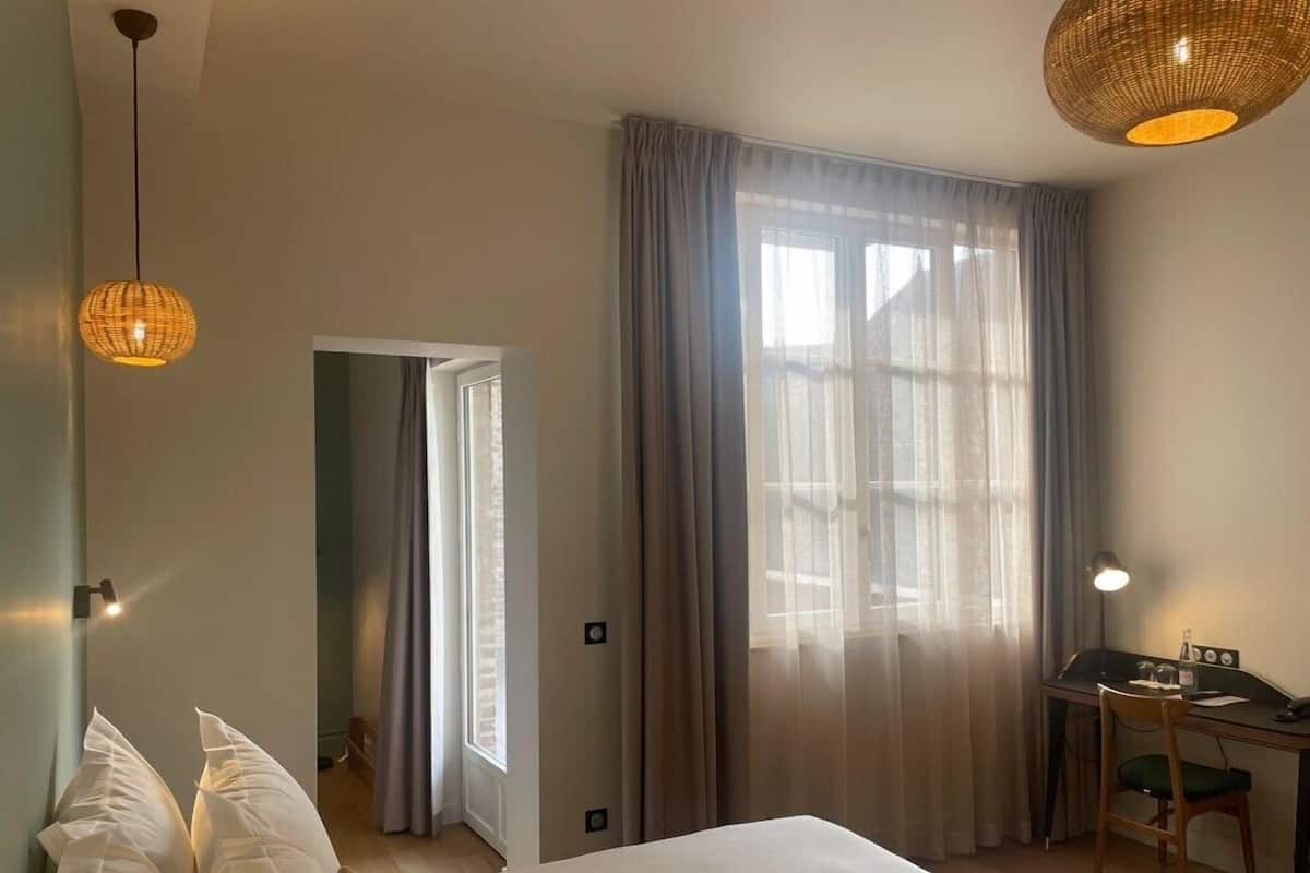 A bedroom with a bed, a desk, and a chair. There is a window with curtains, a door, and a hanging light fixture.