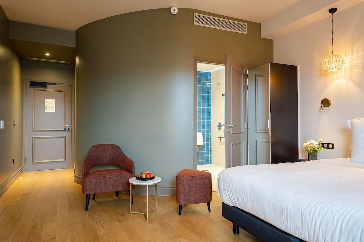 A modern hotel room with a bed, two armchairs, a small table with fruit, and a bathroom visible through an open door.
