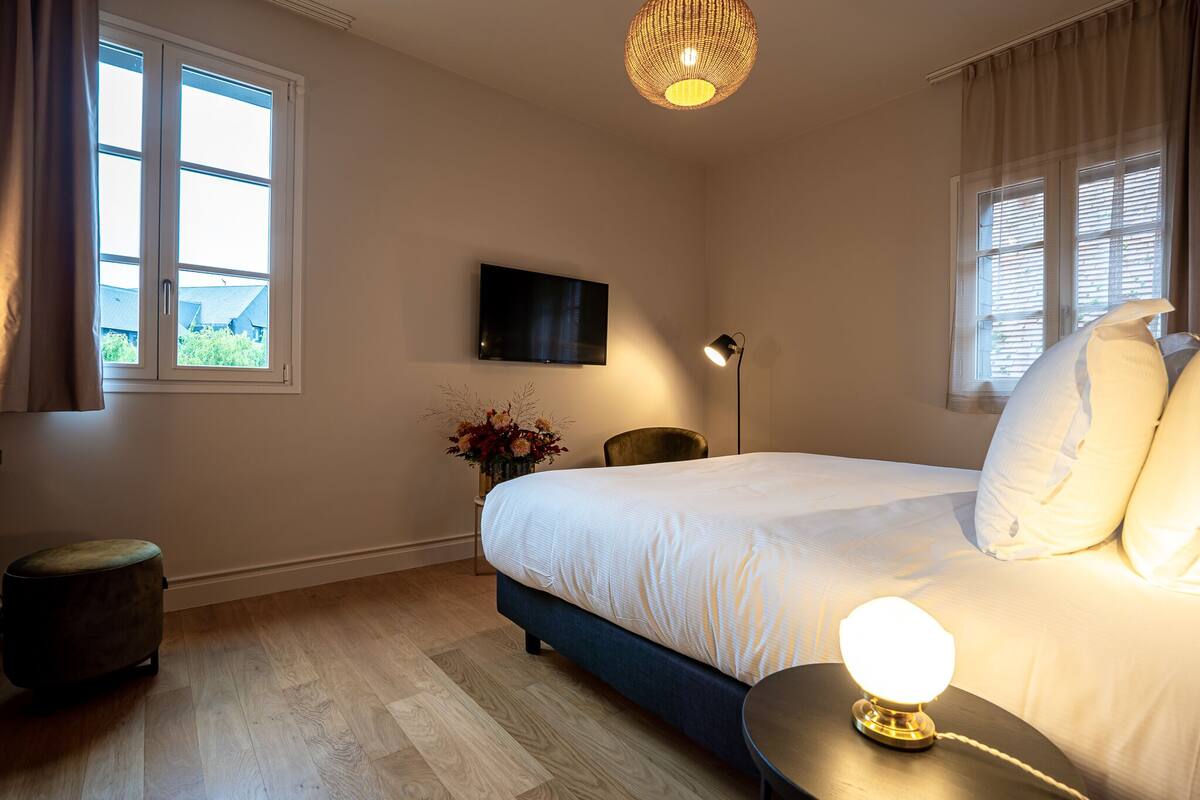 A bedroom with a bed, a TV mounted on the wall, a bedside lamp, and a window with curtains.