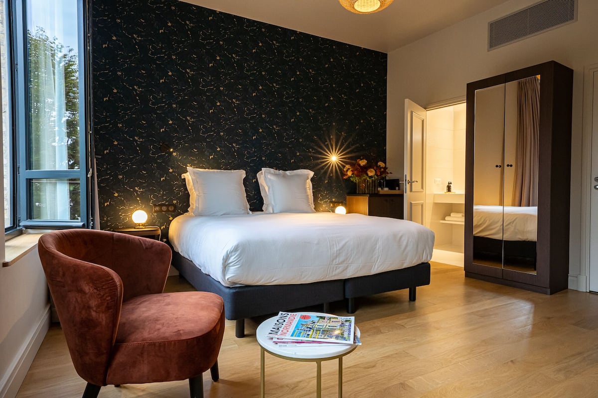 A modern bedroom with a large bed, a nightstand, a chair, a small table with a magazine, and a wardrobe.