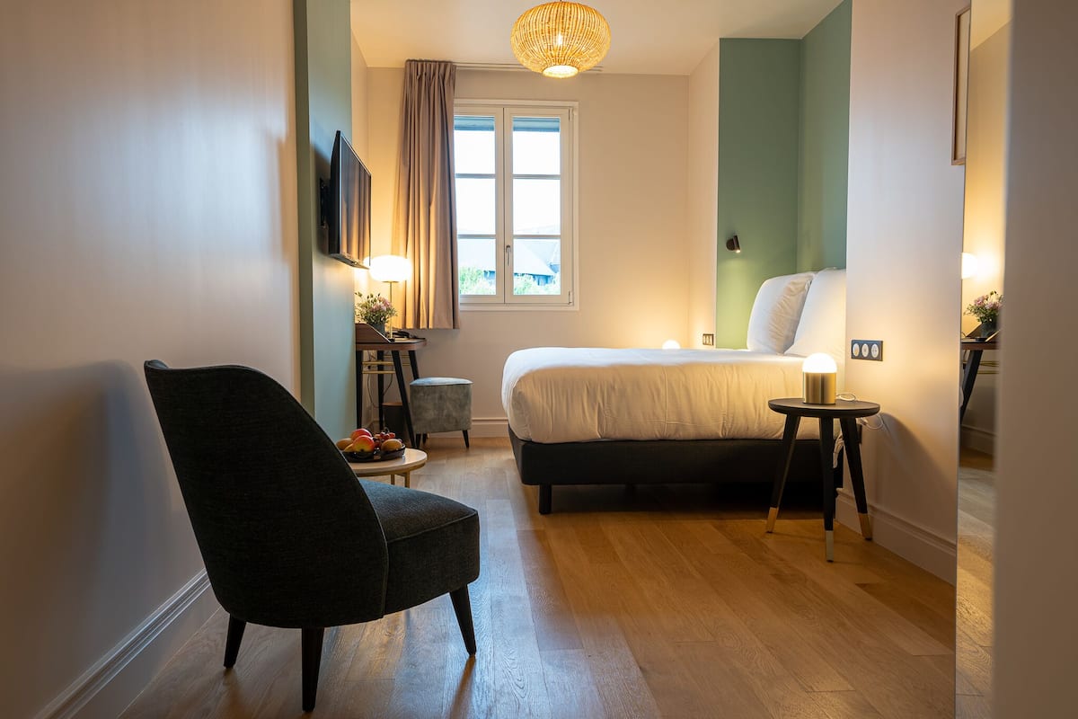 A hotel room with a bed, a chair, a small table with fruit, a TV, and a window with curtains.