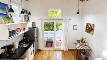 A spacious room with a wooden ceiling and a ceiling fan, a kitchen area with modern appliances, a dining table with chairs, and a large window providing natural light.