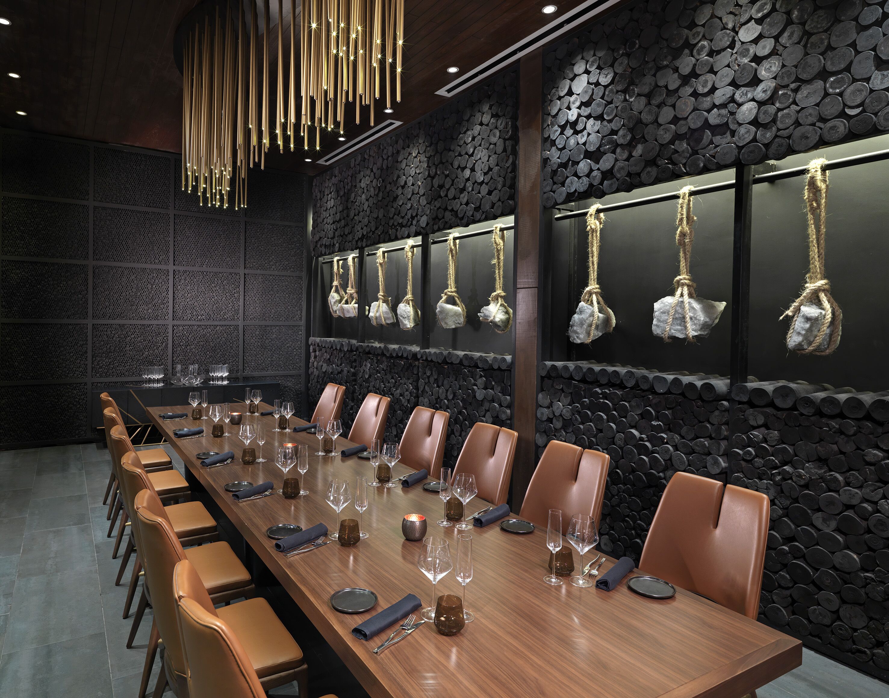Una larga mesa de comedor dispuesta con sillas, vasos y cubiertos, con una pared de troncos y piedras colgantes al fondo.