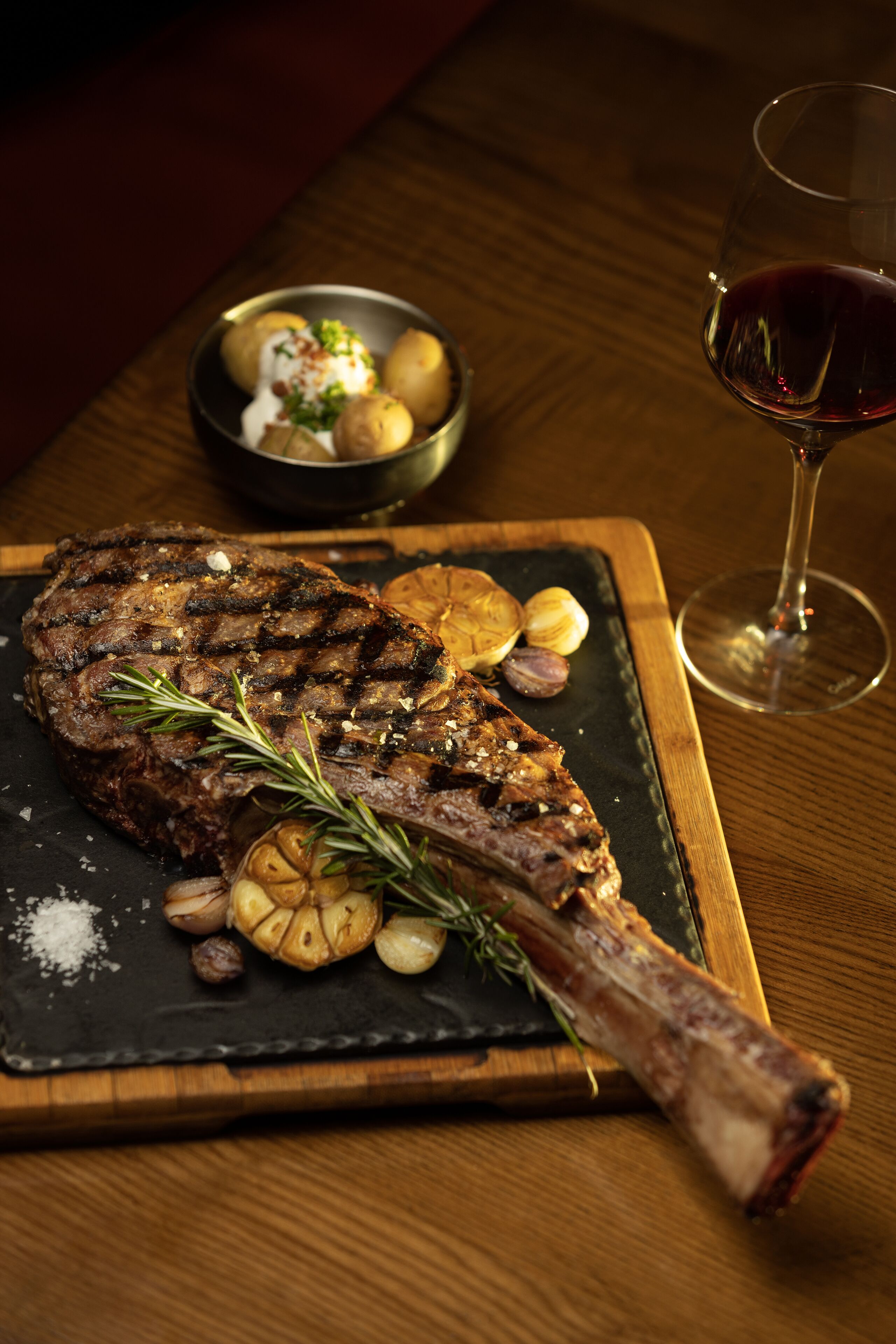 A steak with garlic and rosemary on a wooden board, accompanied by a bowl of potatoes and a glass of red wine.