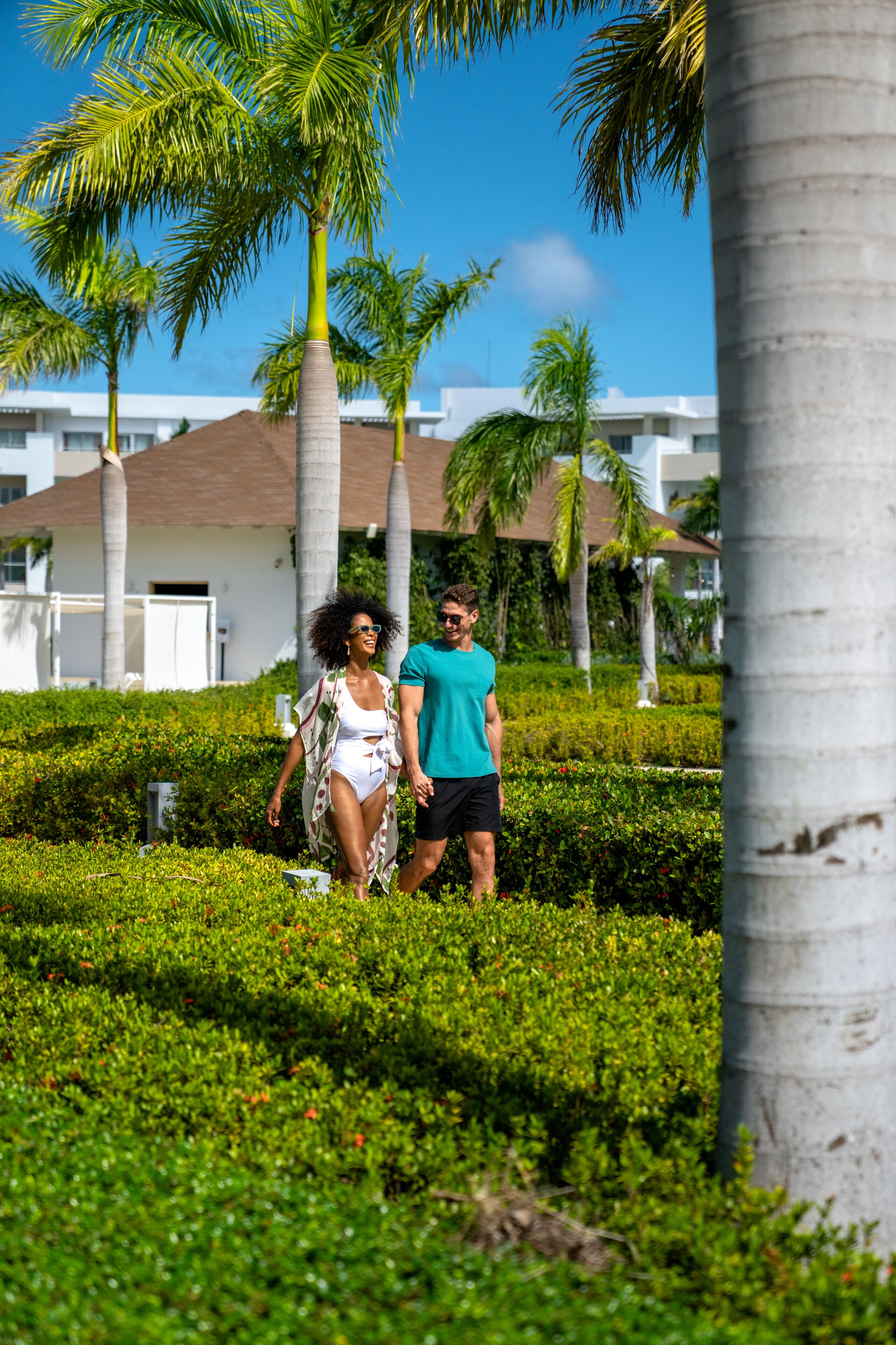 A couple walking on a manicured lawn with palm trees and a building in the background.