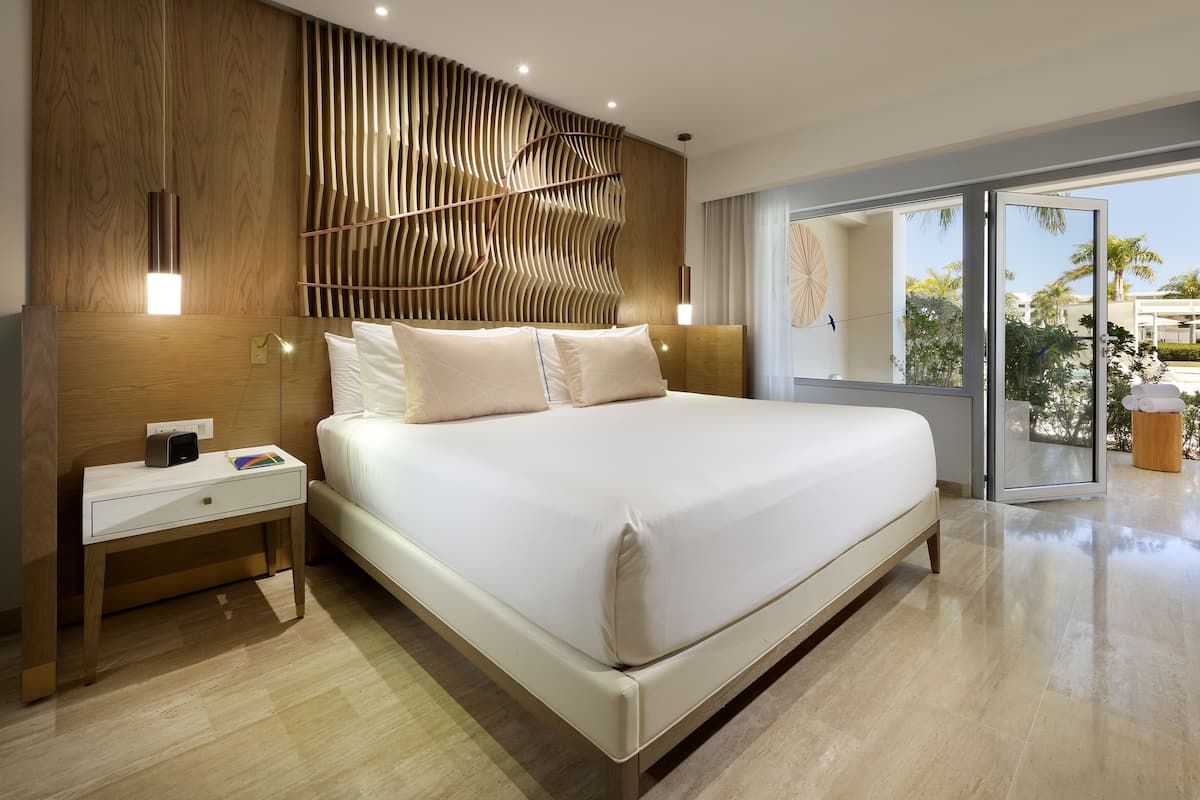 A modern hotel room with a large bed, a bedside table, and a view of palm trees through a glass door.