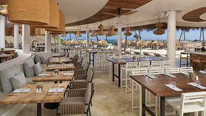 An outdoor dining area with wooden tables and white chairs, surrounded by thatched umbrellas and palm trees, overlooking the beach. - Paradisus Grand Cana - All Suites - All Inclusive (Punta Cana)