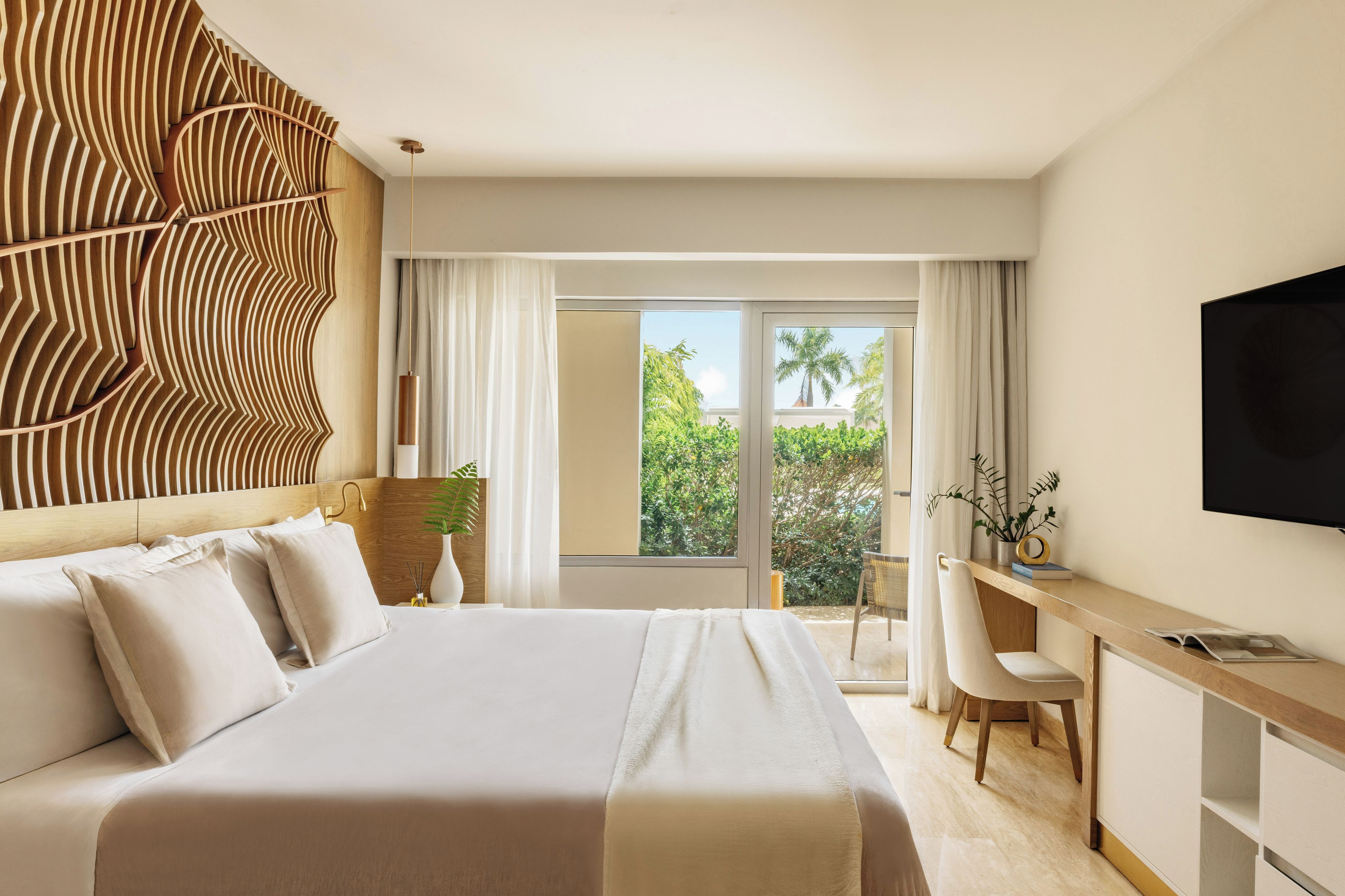 A hotel room with a large bed, a desk, a chair, a TV, and a view of greenery outside.