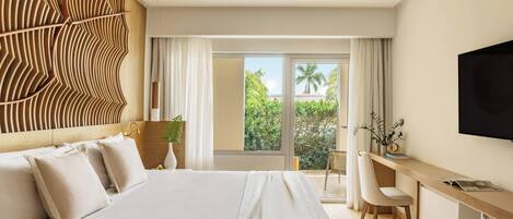 A hotel room with a large bed, a desk, a chair, a TV, and a view of greenery outside.