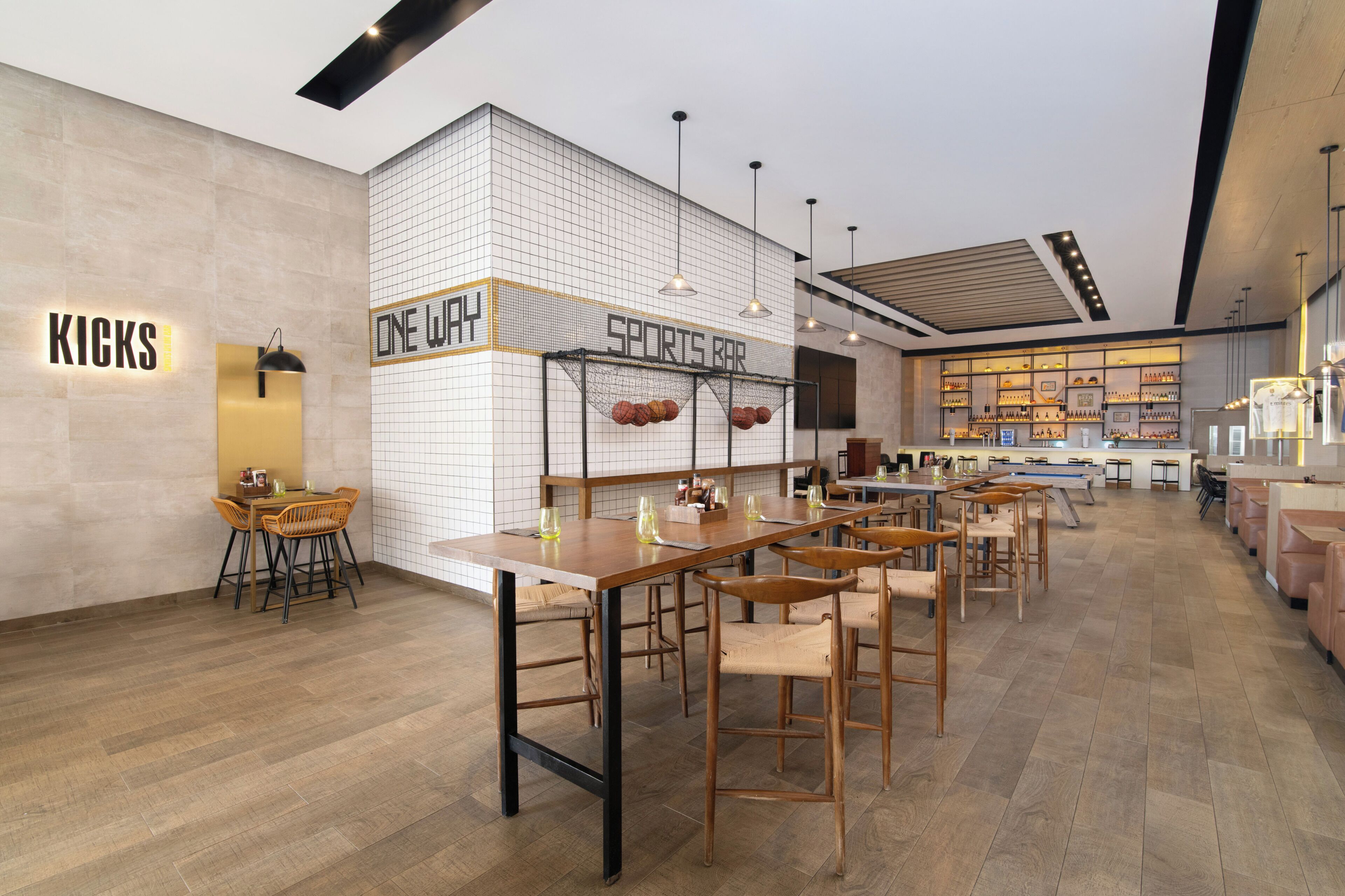 Un bar sportif avec un mur recouvert de carreaux, des tables et des chaises en bois, et une enseigne bien visible sur laquelle on peut lire « KICKS ».