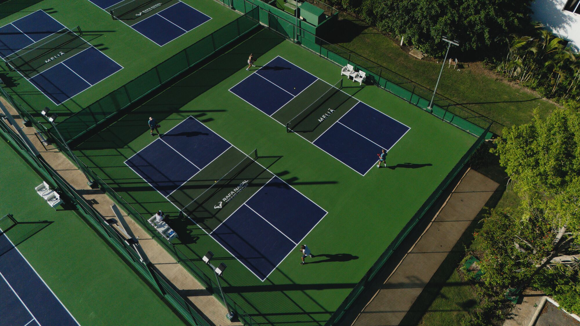 An aerial view of a tennis facility with multiple courts, seating areas, and a surrounding fence.