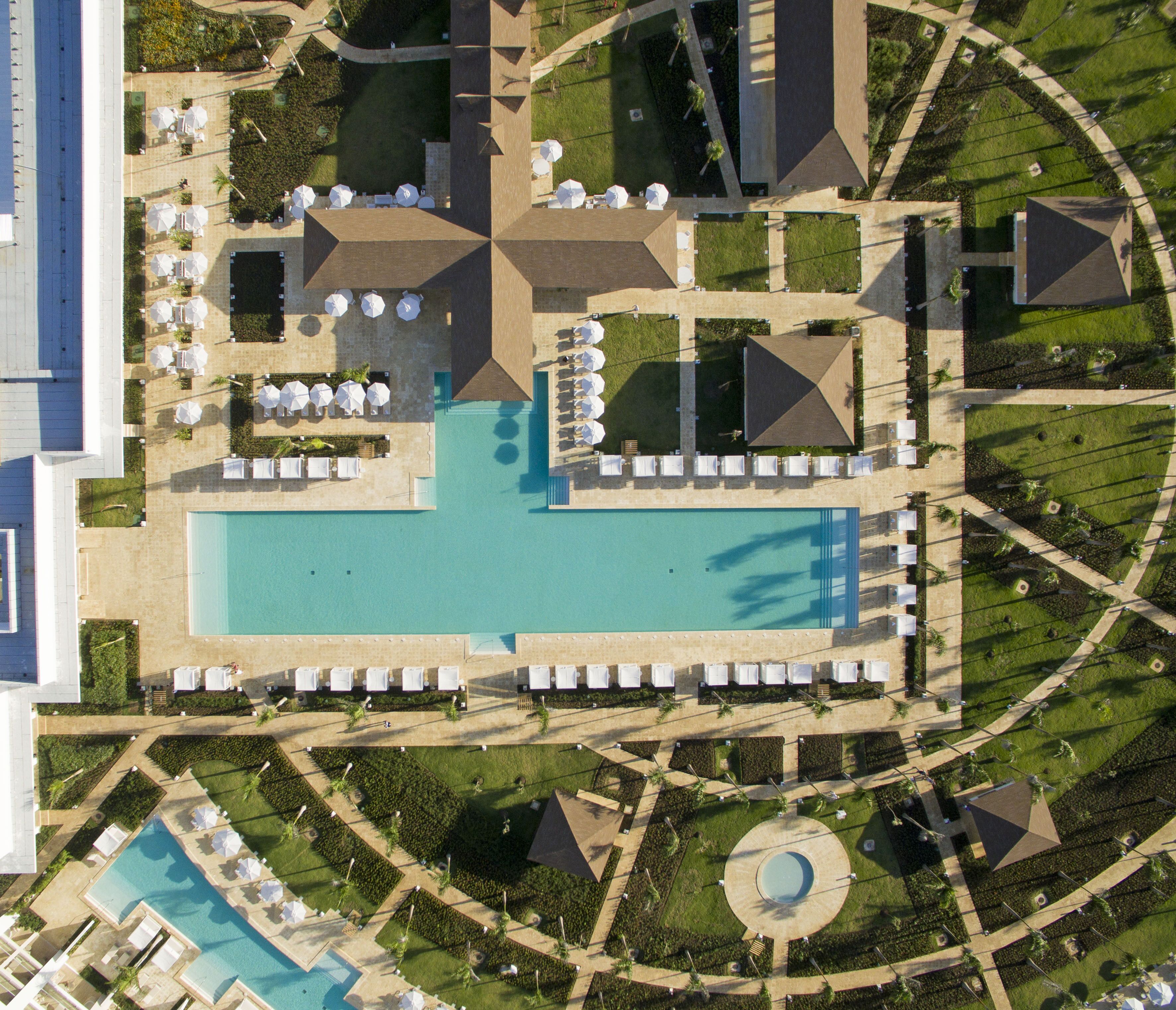 Vista aérea de un resort con una piscina central, varias zonas de descanso con sombrillas y jardines ajardinados.