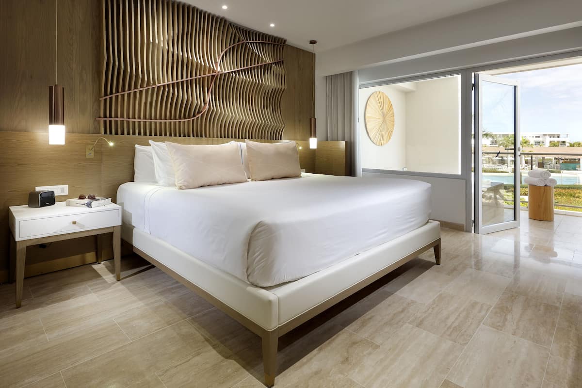 A modern bedroom with a large bed, a bedside table, a wall-mounted light fixture, and a view of a pool through a glass door.