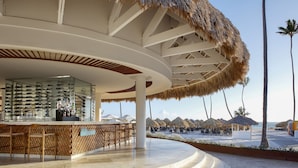 A modern bar area with a curved ceiling, a glass display case of shoes, and a view of the beach with palm trees. - Paradisus Grand Cana - All Suites - All Inclusive (Punta Cana)