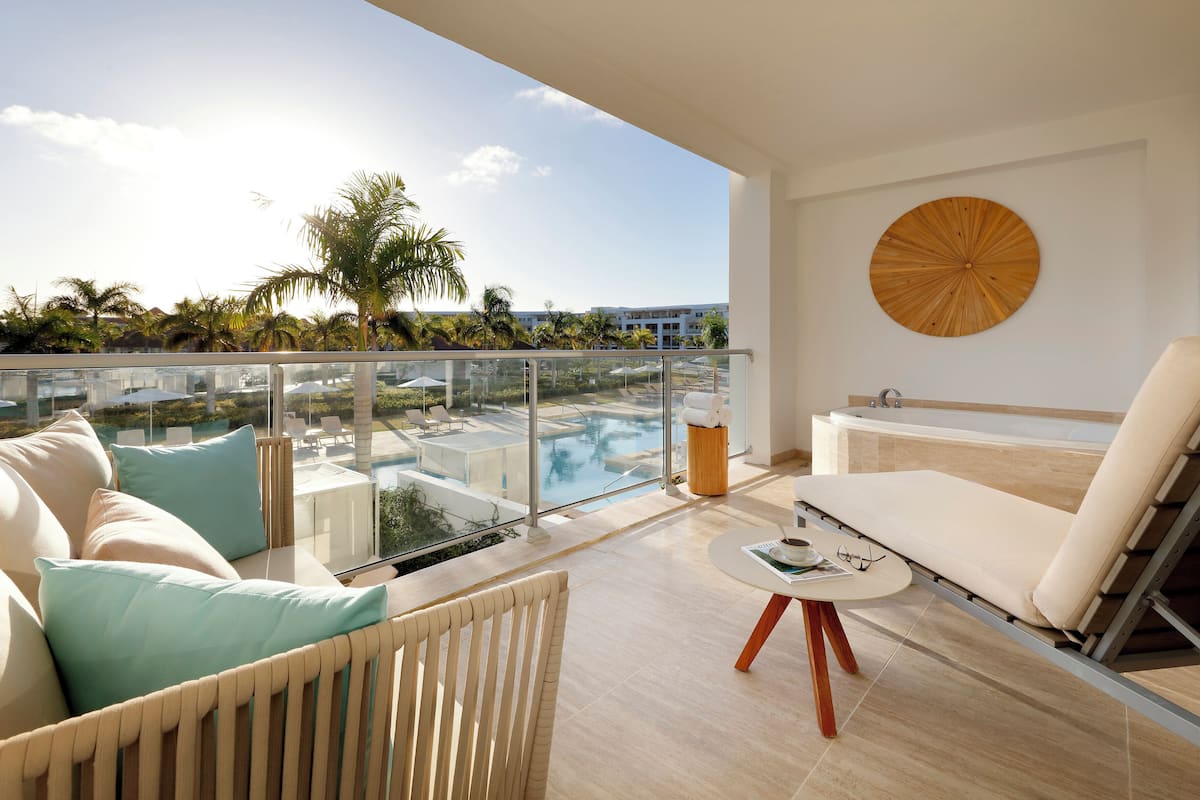 A balcony with a view of a pool and palm trees, a white sofa with cushions, a round wooden table, and a white chair.