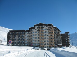A multi-story hotel with balconies, a snow-covered driveway, and a mountainous backdrop. - travelski home classic - Résidence Médian - Studio 4 personnes 308 (Les Menuires)